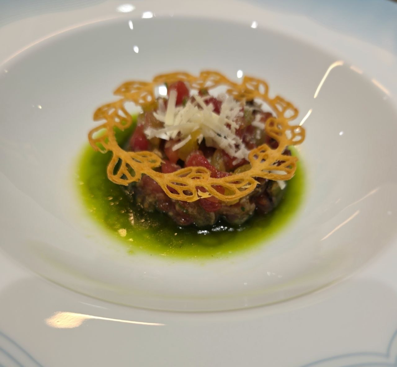 Tomato tartare with shaved Parmesan and a lace tuile, in a basil and cucumber juice, in a wide white bowl.