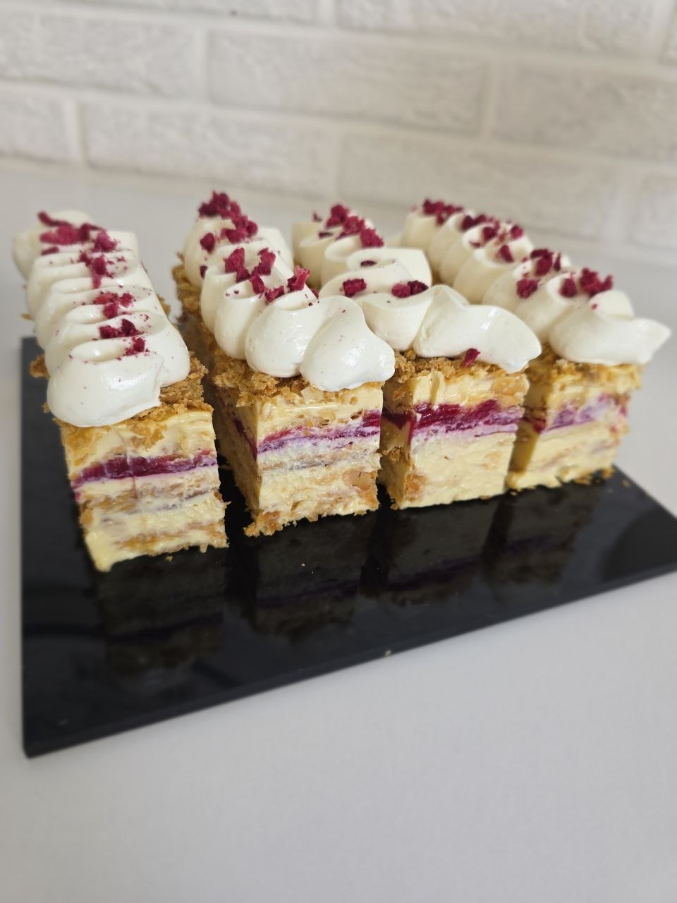 Portions of Napoleon cake showing layers of puff pastry, custard cream and raspberry confit, topped with piped cream and freeze-dried raspberry, on a black slate.