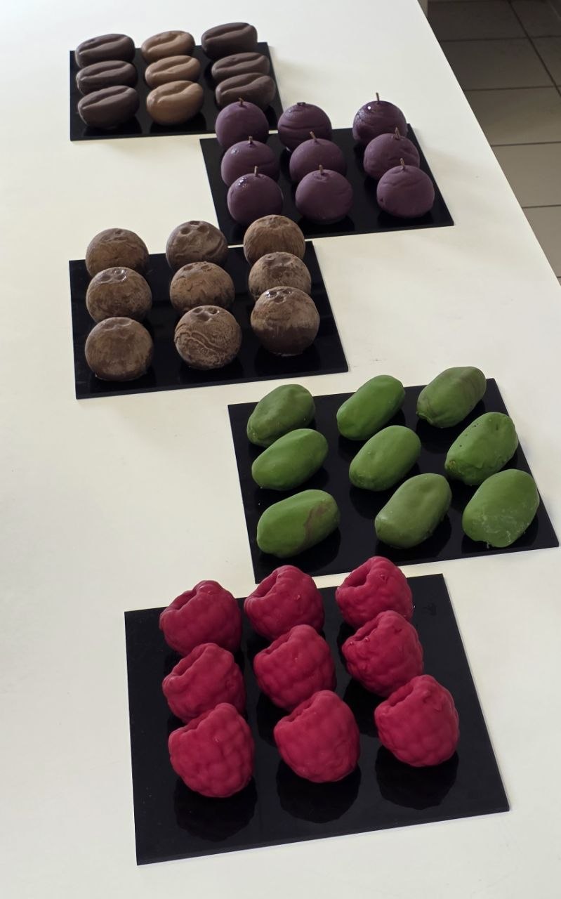 Fruit-shaped mousse desserts finished with a velvet spray, in raspberry and pistachio, on black plates.